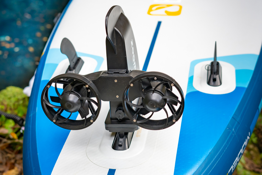  Close-up of a motorized fin attachment on a paddleboard, featuring two black fan-like motors and a sleek blue design.